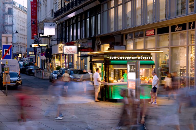 Bustling evening street view of Vienna with people passing by a food stand.